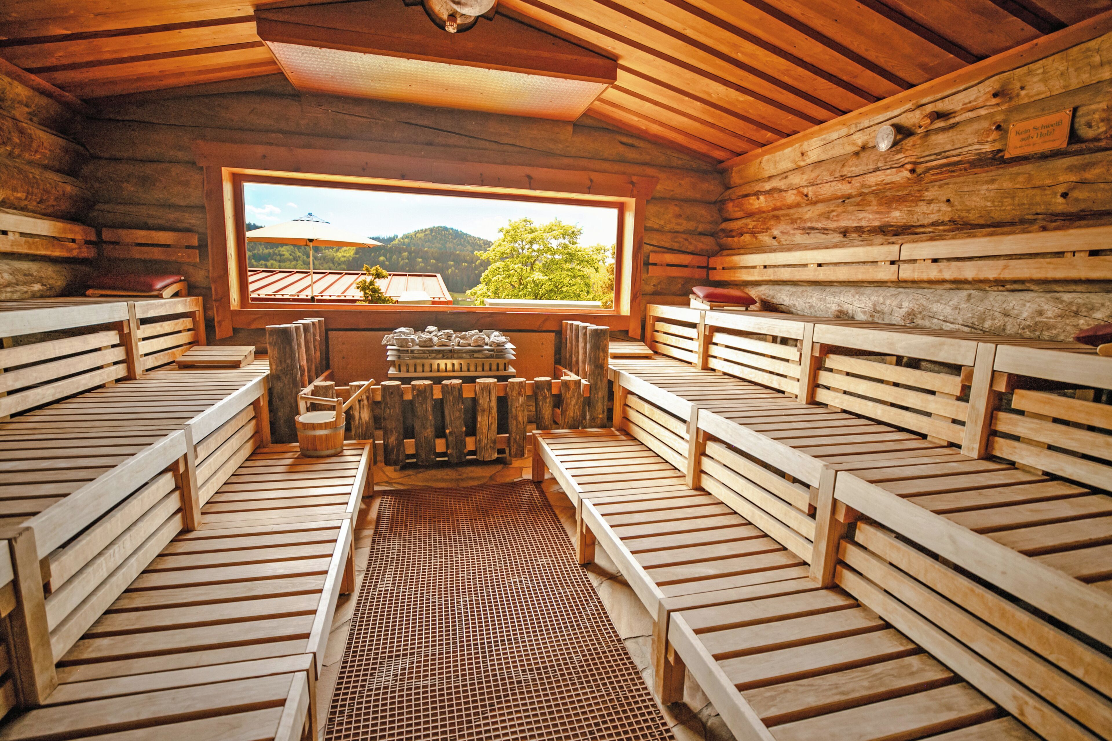 A sauna room with wooden benches, a central heating unit, and a window offering a view of a forested landscape.