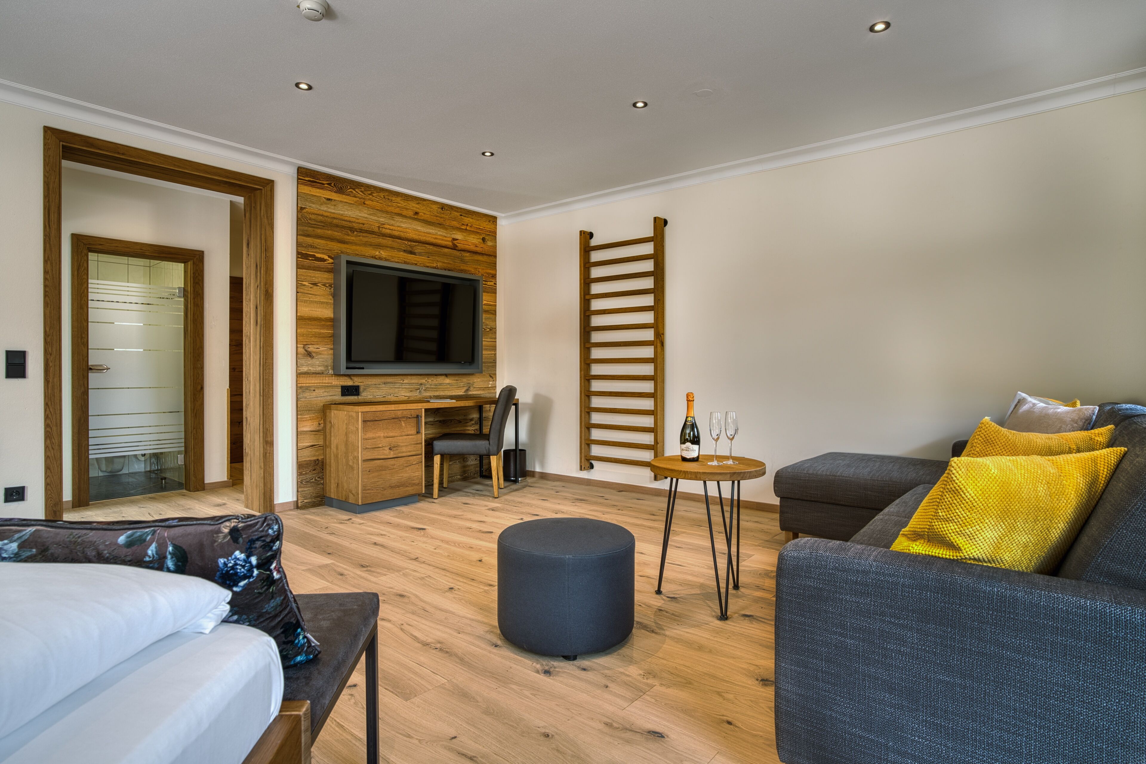 Habitación de hotel moderna con suelo de madera, una cama, un sofá, un televisor montado en la pared y una mesita con una botella y vasos.