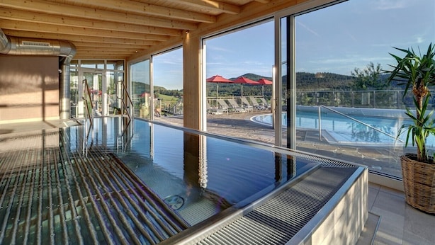 A modern indoor swimming pool with a glass barrier, wooden ceiling, and a view of a beach and mountains. - Wellnesshotel Hofbräuhaus (Bodenmais)