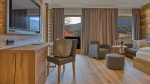 A modern hotel room with a wooden desk, a comfortable seating area, and a large window offering a scenic mountain view. - Wellnesshotel Hofbräuhaus (Bodenmais)