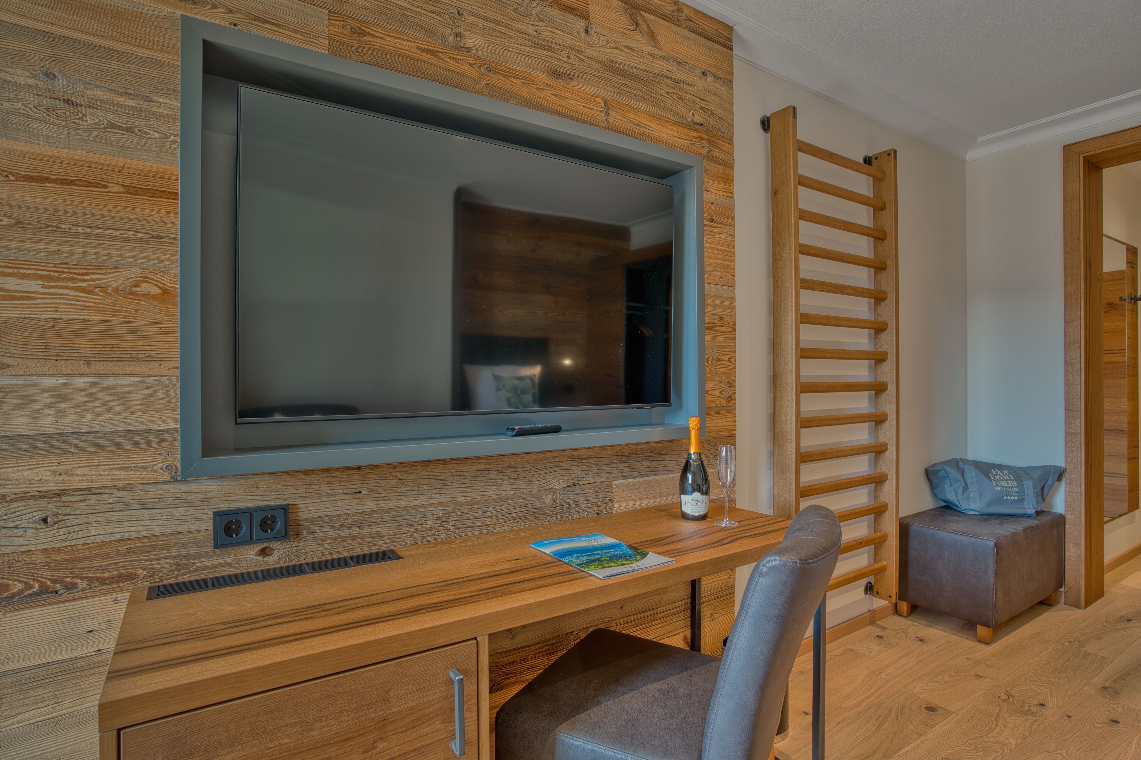 Una habitación moderna con un televisor de pantalla plana grande montado en una pared de madera, un escritorio de madera con una silla, un taburete pequeño y una botella de champán.
