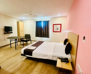 A bedroom with a bed, a desk, a chair, a TV, and a window with curtains. - Hotel Royal Address (Noida)