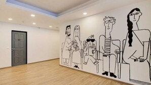 Een kamer met een houten vloer, een deur en een muur met een muurschildering van zittende figuren.
