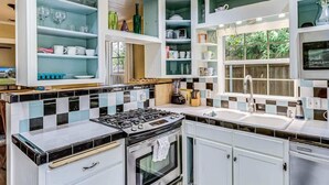 Une cuisine équipée d’une cuisinière à gaz, d’un four et de meubles de rangement blancs. On y trouve des étagères remplies d’assiettes et de verres.