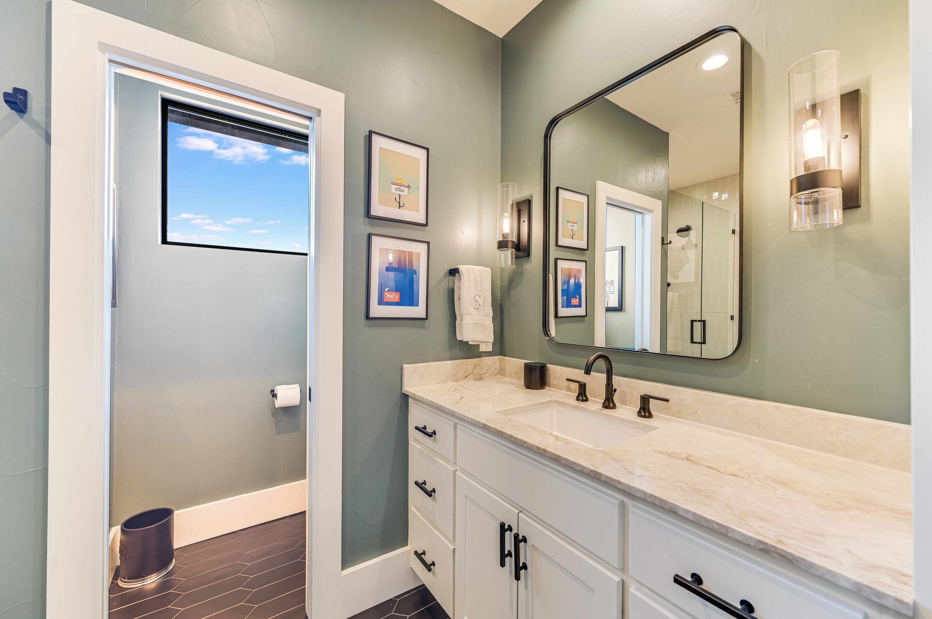 Une salle de bain avec un grand miroir, un lavabo et des toilettes. Des tableaux encadrés ornent le mur et une serviette est accrochée à la porte.