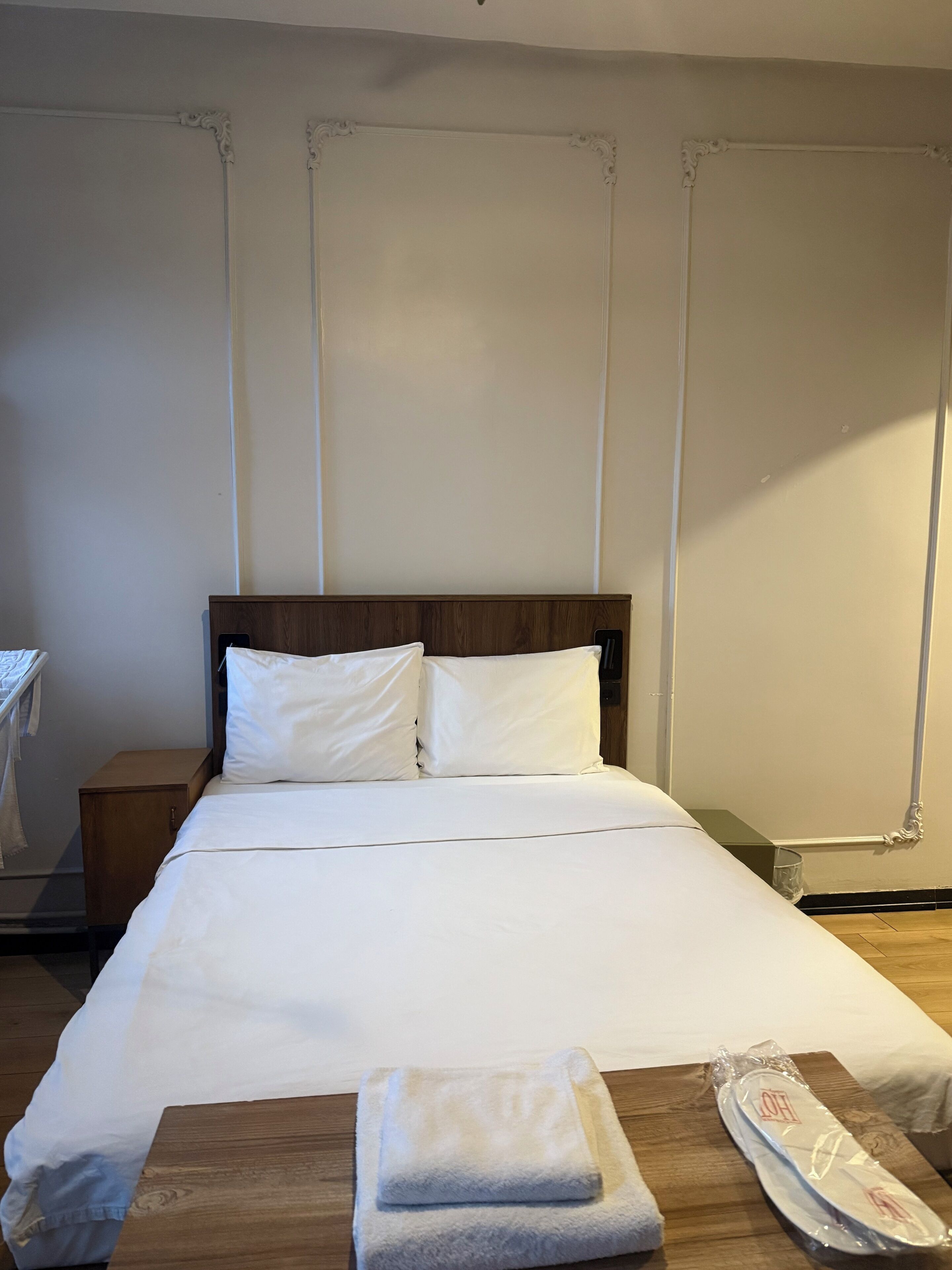 A neatly made bed with white linens, a wooden nightstand, and a small table with a folded towel and a tissue box.