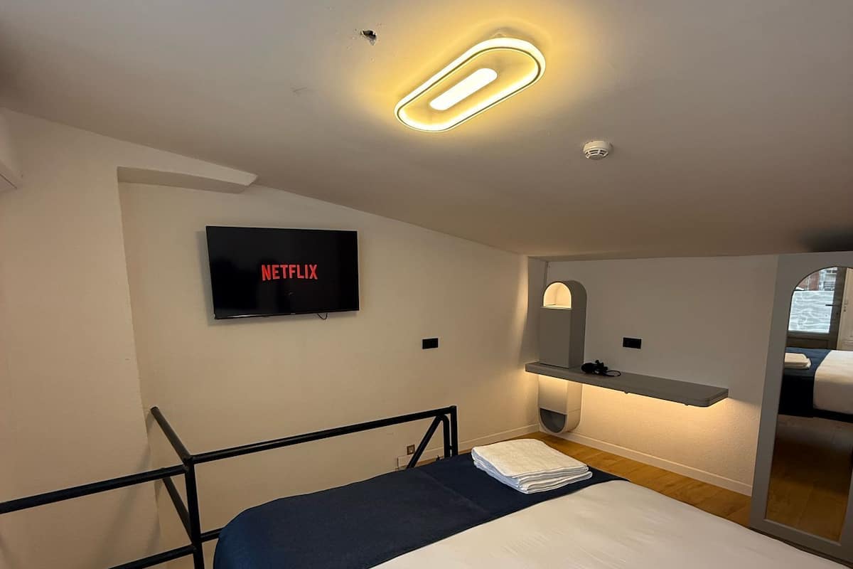 A hotel room with a bed, a TV mounted on the wall, a bedside table with a lamp, and a window with a view.
