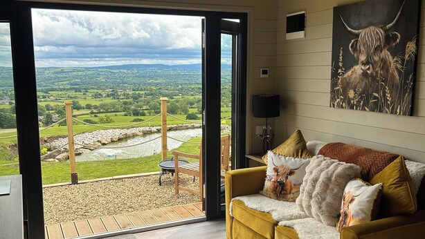 A room with a yellow sofa, a painting of a bull, and a view of a green landscape through a glass door. - The Highland Cow Bothy (Longridge)