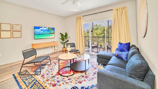 A living room with a sofa, chairs, a TV, a ceiling fan, a balcony with curtains, and a large window. - Family-friendly Condo With Pool, Sauna & Hot Tub (Winter Garden)