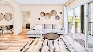 Uma sala de estar moderna com um sofá seccional, uma mesa de centro redonda e um espaço de jantar com mesa e cadeiras. Há uma varanda com porta de vidro e ventilador de teto.