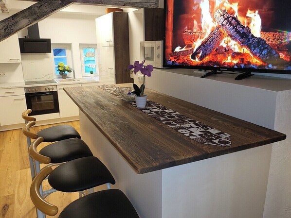 A modern kitchen with a wooden bar counter, a television displaying a fire, and a bar stool seating arrangement.