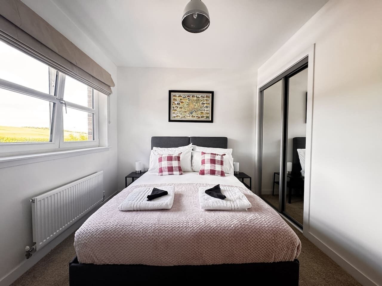 Een net opgemaakt bed met een roze sprei, twee kussens en twee opgevouwen handdoeken. Daarboven hangt een lijstje op de muur, en er is een raam met uitzicht op de omgeving. Een radiator is aan de muur bevestigd.