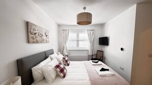 A bedroom with a bed, a nightstand, a lamp, a window with curtains, a wall-mounted TV, and a radiator.