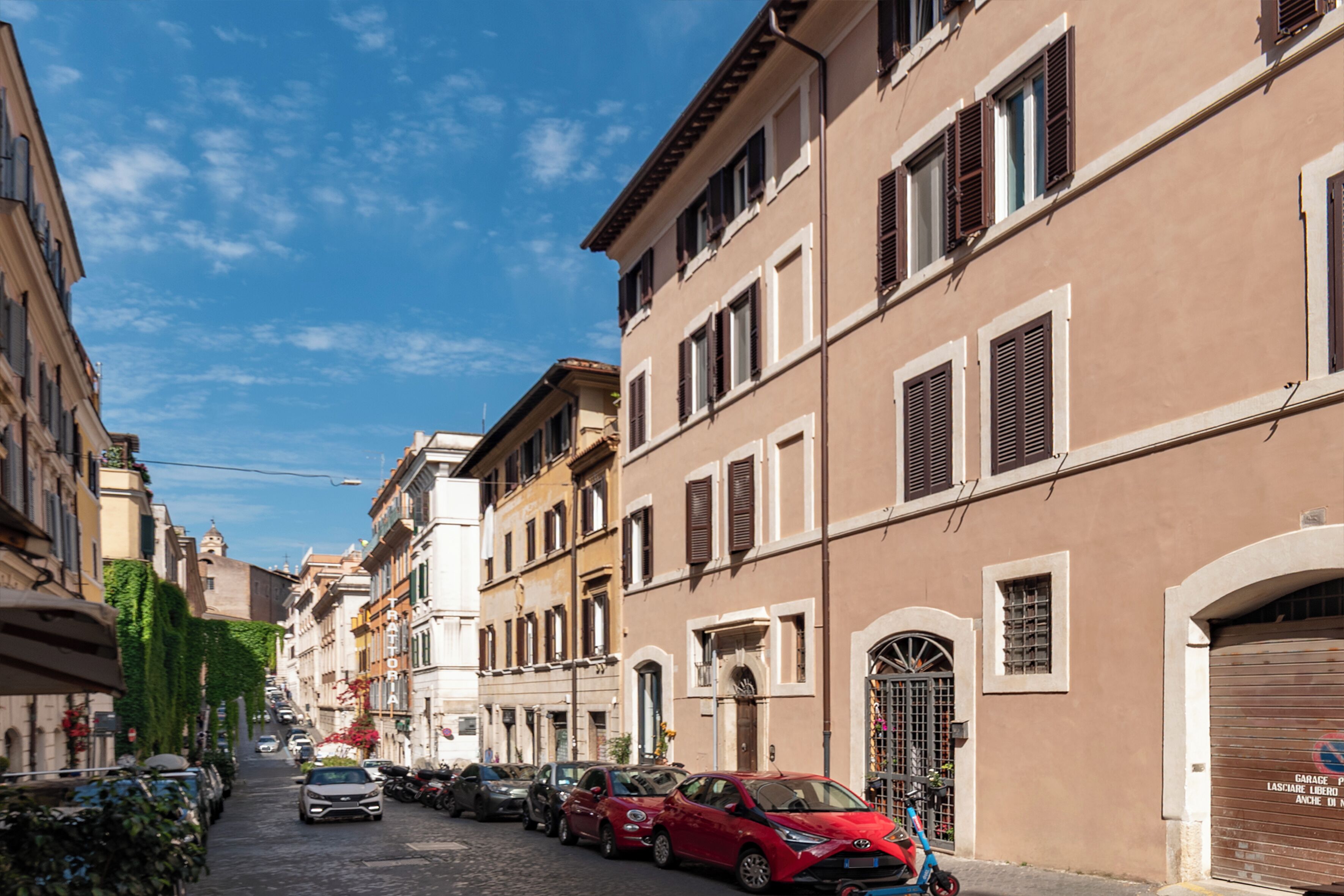 A narrow cobblestone street lined with parked cars and historic buildings with arched doorways and shutters.