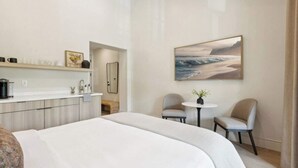 A modern hotel room with a large bed, a small round table, two chairs, a kitchenette, and a painting of a beach scene. - Casa Riviera 29 - Boutique Suites (Santa Barbara)