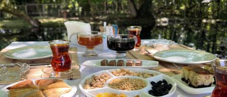 An outdoor dining table with various dishes, including pastries, nuts, and a bowl of salad, set against a backdrop of trees.