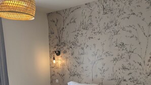 Uma cama bem arrumada com roupa de cama branca e um manta verde, uma luminária de parede e um papel de parede com padrão floral.