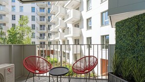 A balcony with two red chairs and a small table, a green hedge, and a view of adjacent buildings. - Żurawie IKONY 1 by Grand Apartments (Gdansk)