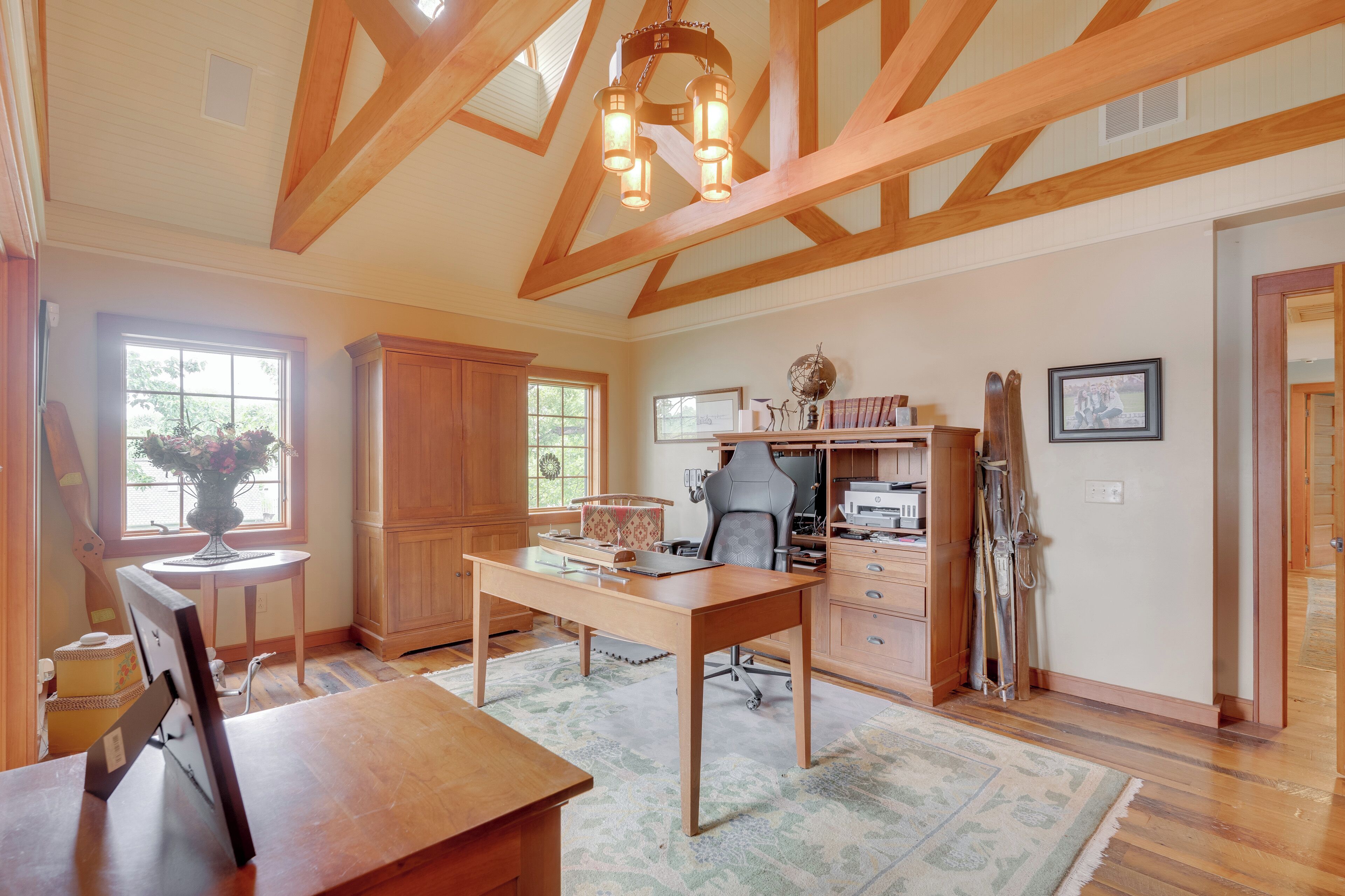 A spacious room with a wooden desk, a chair, a large window, and a ceiling with exposed beams.