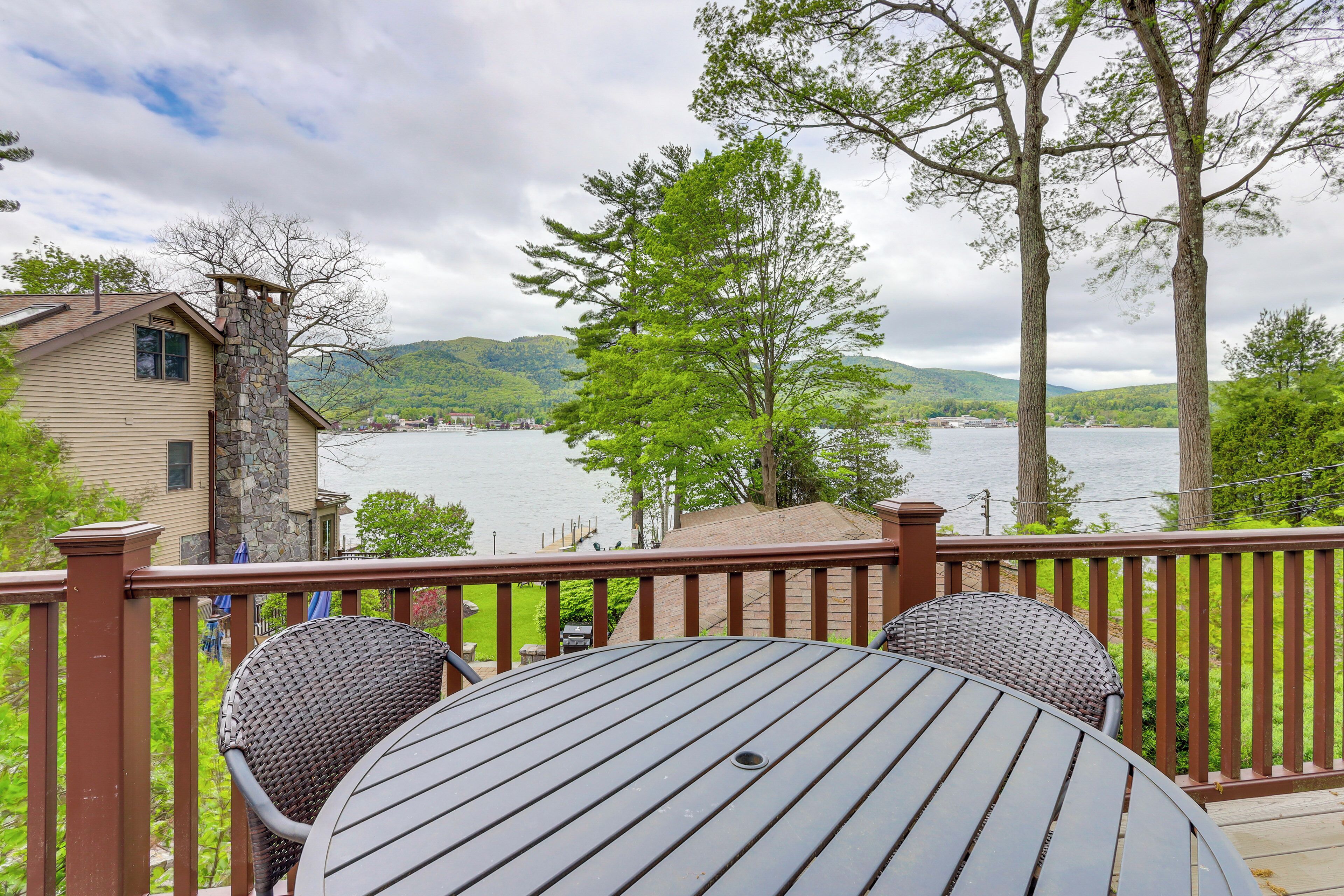 A deck with a table and chairs overlooking a lake, surrounded by trees and a house.