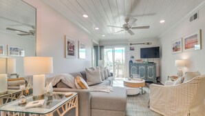 A spacious living room with a large sofa, a glass coffee table, and a television. There is a ceiling fan, a fireplace, and a view of the outdoors. - Walk to Beach & Marsh: Condo in Murrells Inlet (Murrells Inlet)