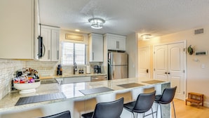 A modern kitchen with white cabinets, a stainless steel refrigerator, a large island with bar stools, and a window with blinds. - Lake & Pool Access: Lazy Days Condo w/ Balcony! (Osage Beach)
