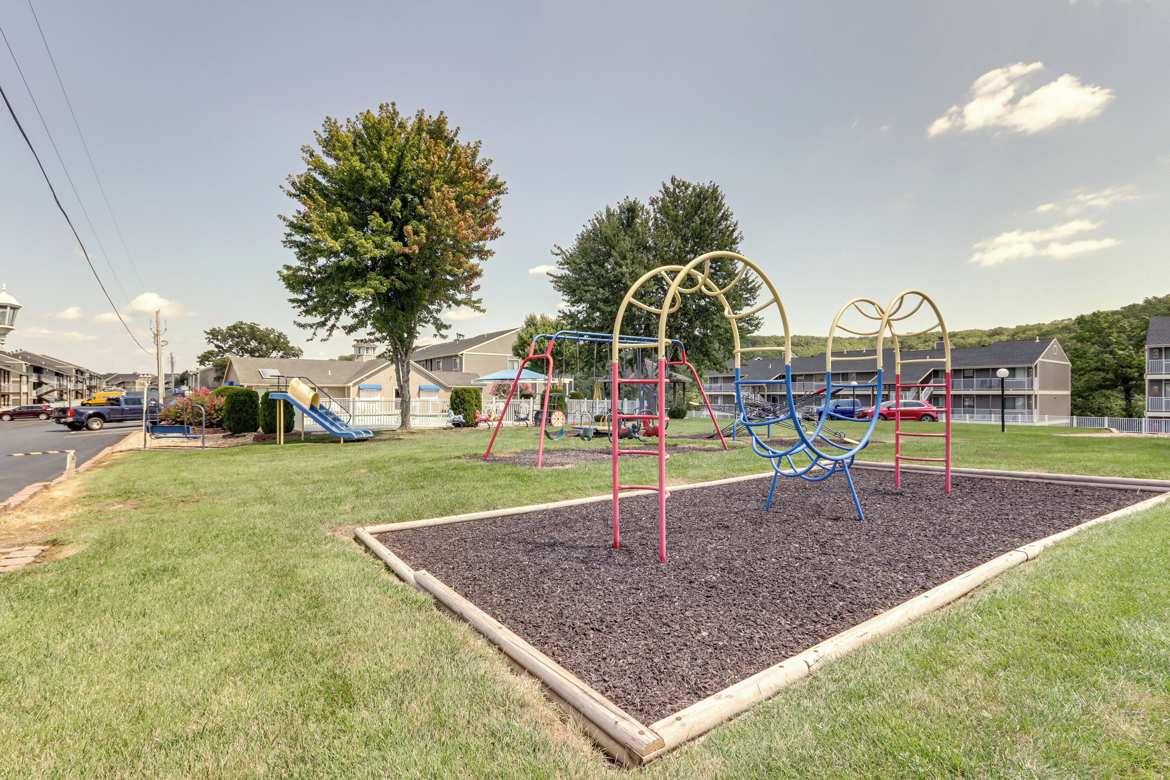 A playground with various play structures, surrounded by a grassy area and residential buildings in the background.