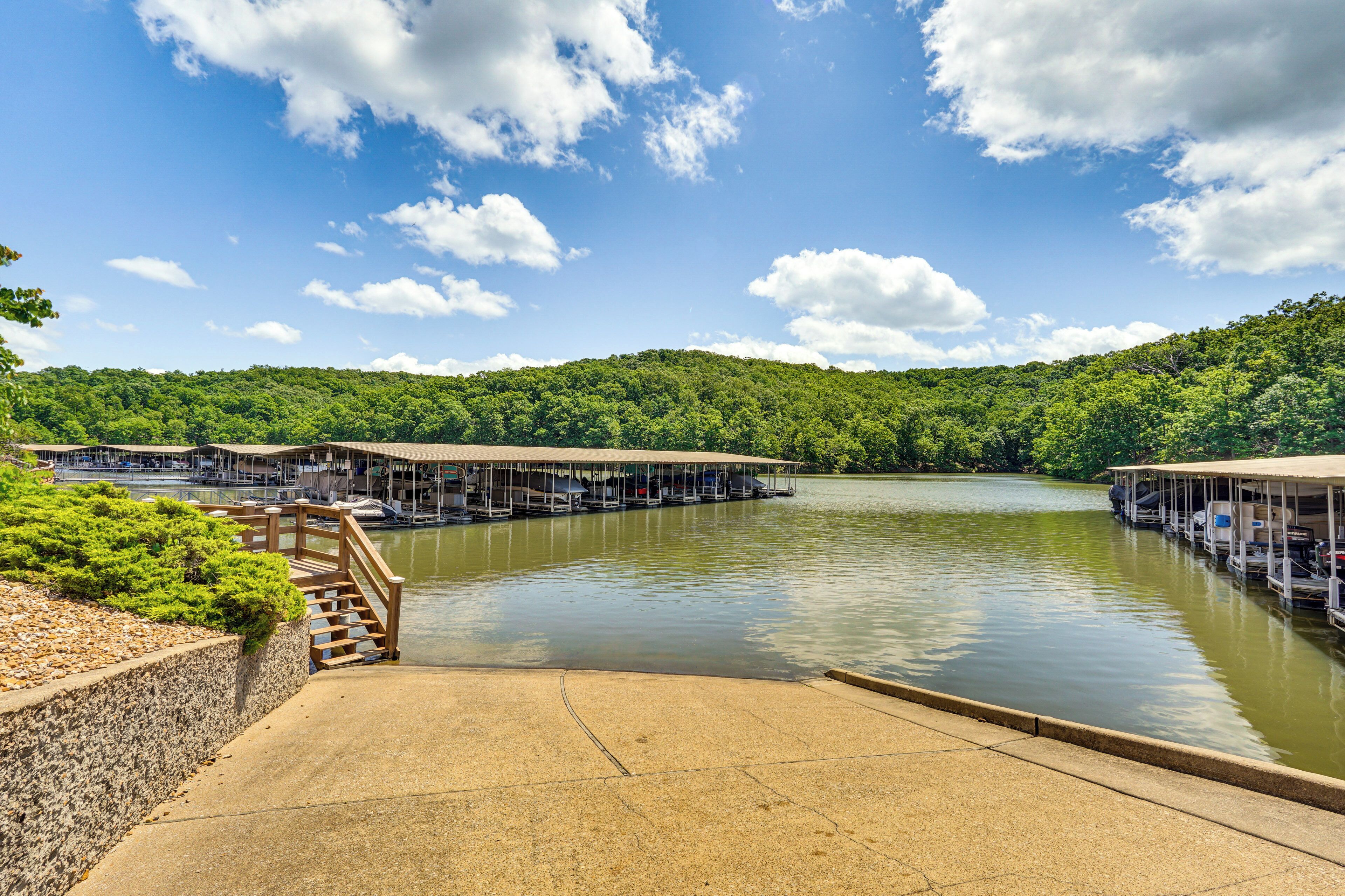 A marina with docked boats, a concrete walkway, and a forested hillside in the background.