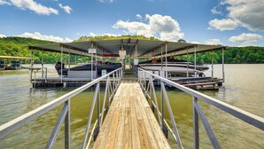A wooden dock leading to a covered boat launch area with a view of a lake and forested hills. - Lake & Pool Access: Lazy Days Condo w/ Balcony! (Osage Beach)