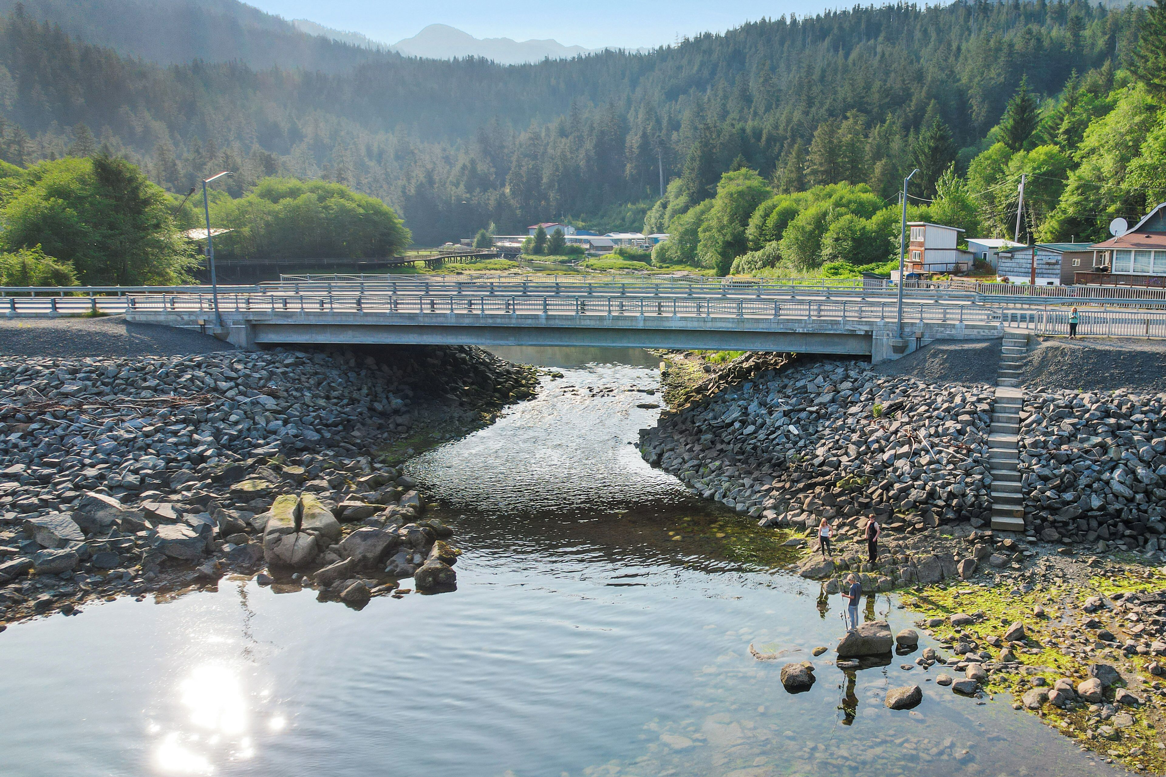 A bridge over a river with a rocky shore, surrounded by greenery and houses.