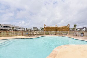Une piscine dotée d’une terrasse en bois et d’un espace salon couvert, le tout entouré d’une clôture en bois et avec des maisons en arrière-plan.