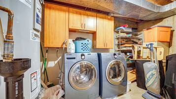 A laundry area with two front-loading washing machines, wooden cabinets, and a shelf with various items.