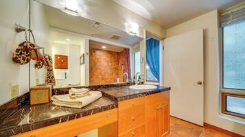 A bathroom with a marble countertop, wooden cabinets, a large mirror, and a view of the outside through a window.