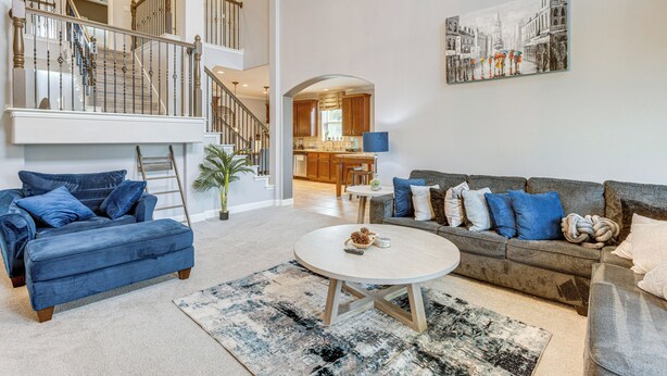 Un salon spacieux avec un canapé bleu, un canapé gris et une table basse blanche. Un escalier mène à un niveau supérieur, et on aperçoit une cuisine au fond.