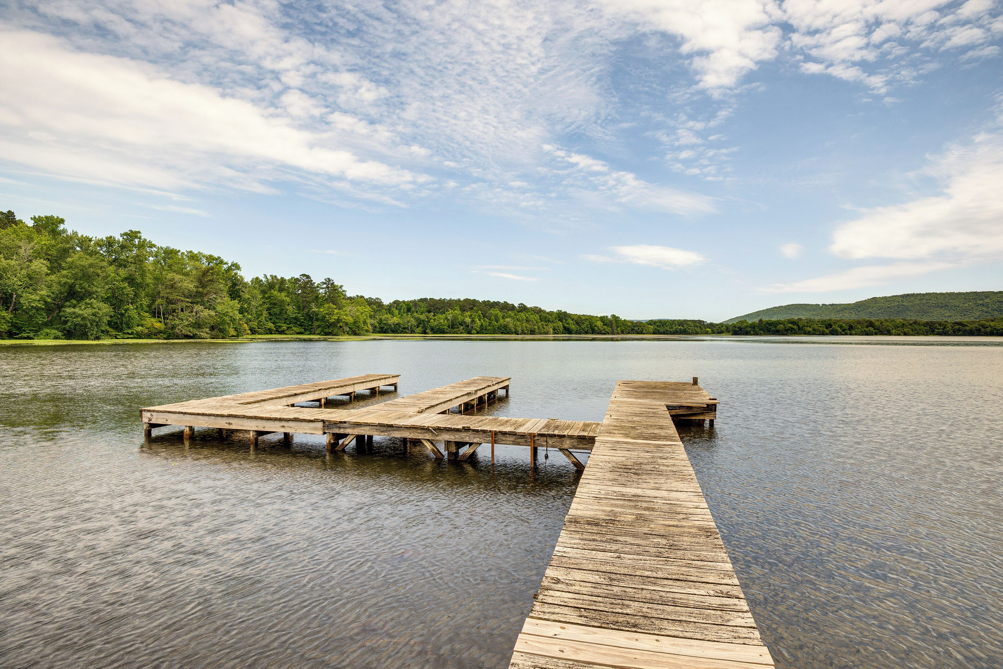 A wooden pier extending into a calm lake, surrounded by dense green trees and hills in the background.