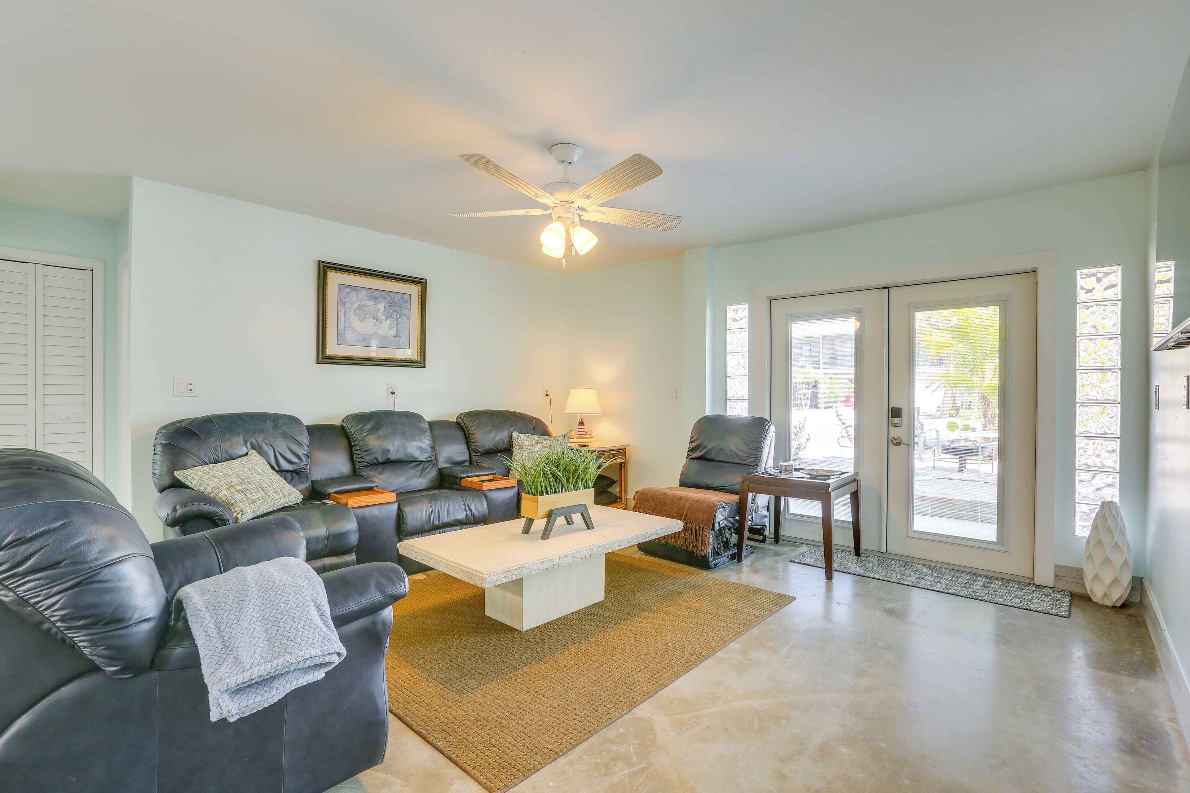 A living room with a sofa, coffee table, and chairs. There is a ceiling fan, a framed picture on the wall, and a door leading outside.