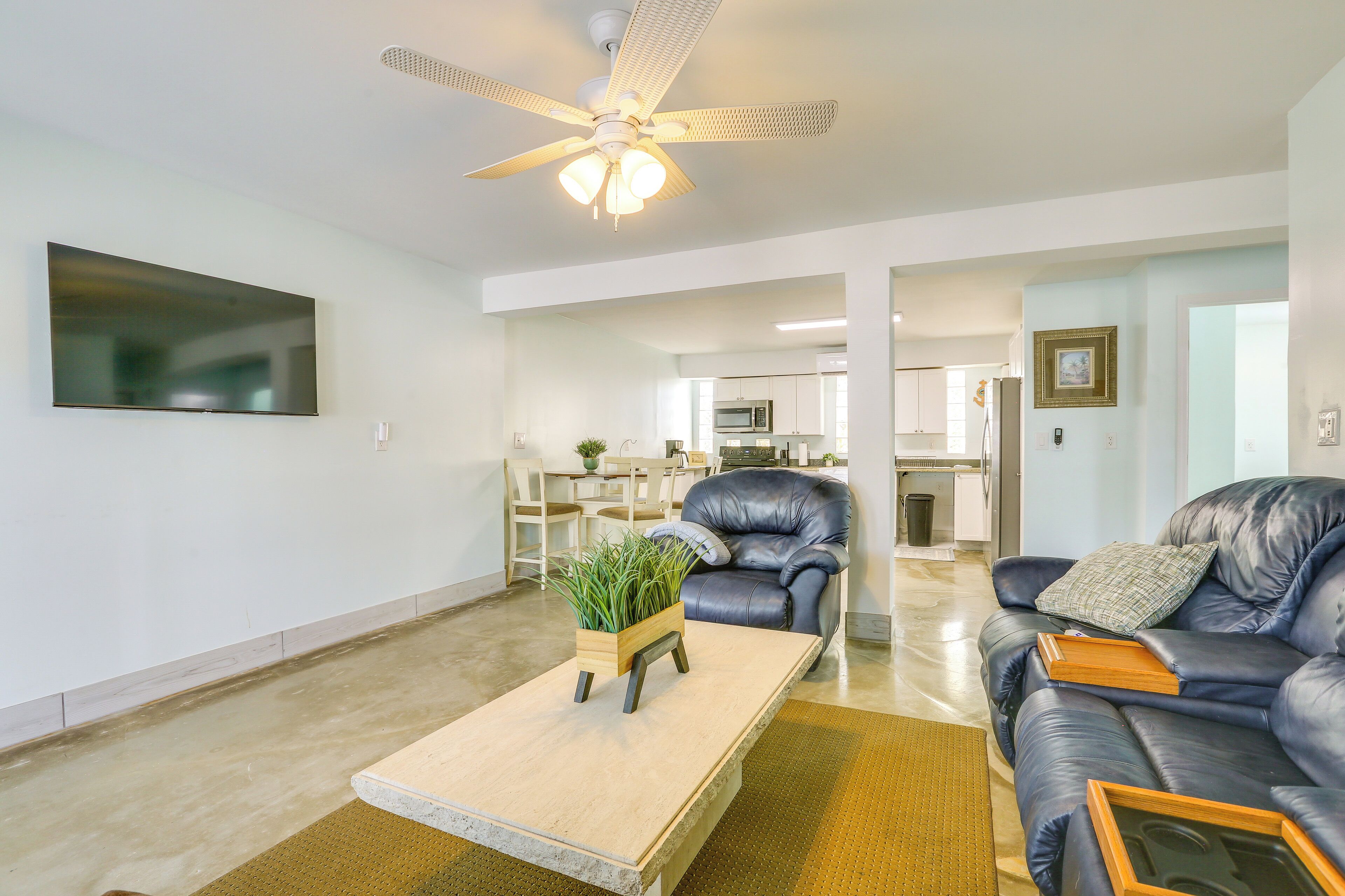 A modern living room with a flat-screen TV, a ceiling fan, a leather sofa, a coffee table with a plant, and a kitchen in the background.
