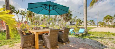 Un patio al aire libre con una mesa de madera y sillas de mimbre, una sombrilla color aguamarina y palmeras al fondo.