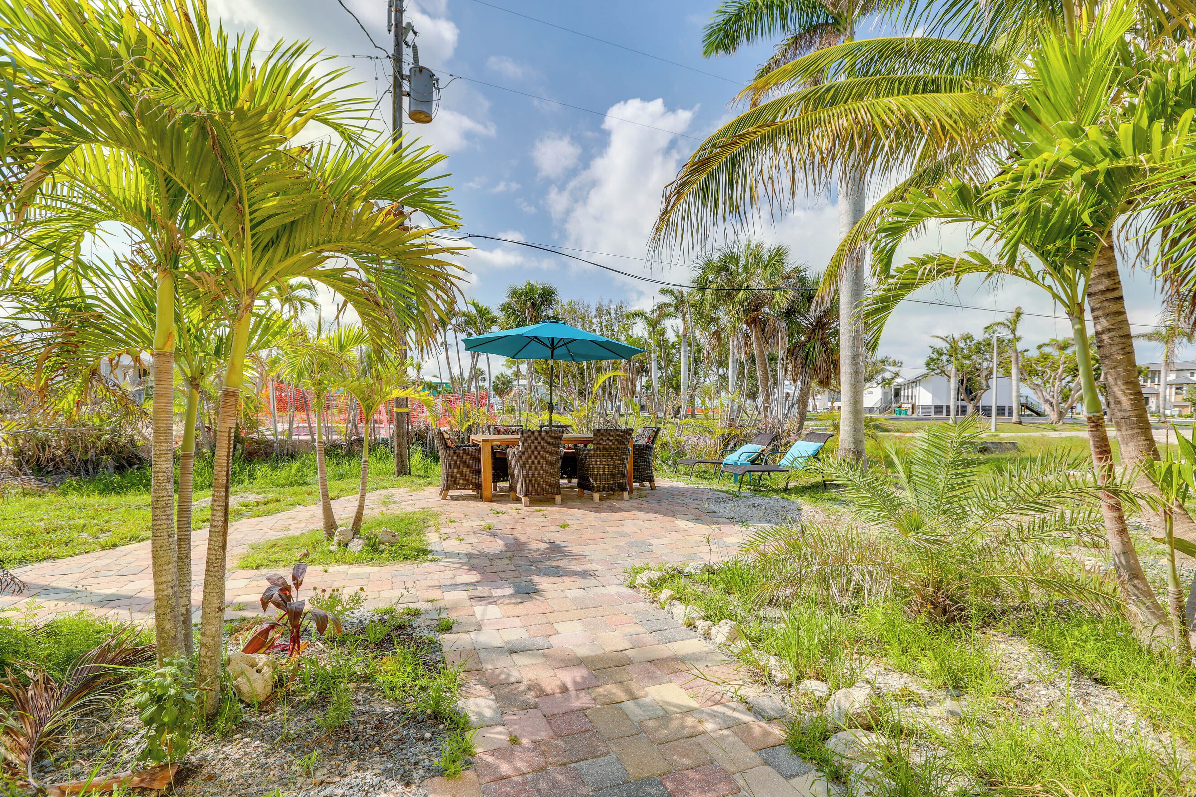 A paved patio with a table and chairs under a blue umbrella, surrounded by palm trees and greenery.