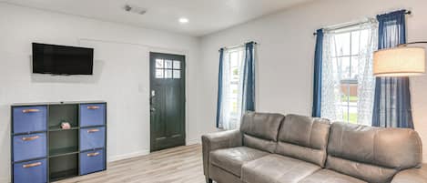 Una sala de estar con un sofá de cuero, un televisor de pantalla plana montado en la pared, un mueble con cajones y una puerta.