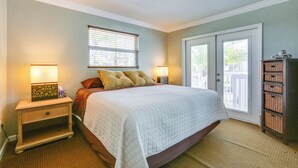 A bedroom with a bed, two bedside tables with lamps, a window with blinds, a door, and a small wooden cabinet.
