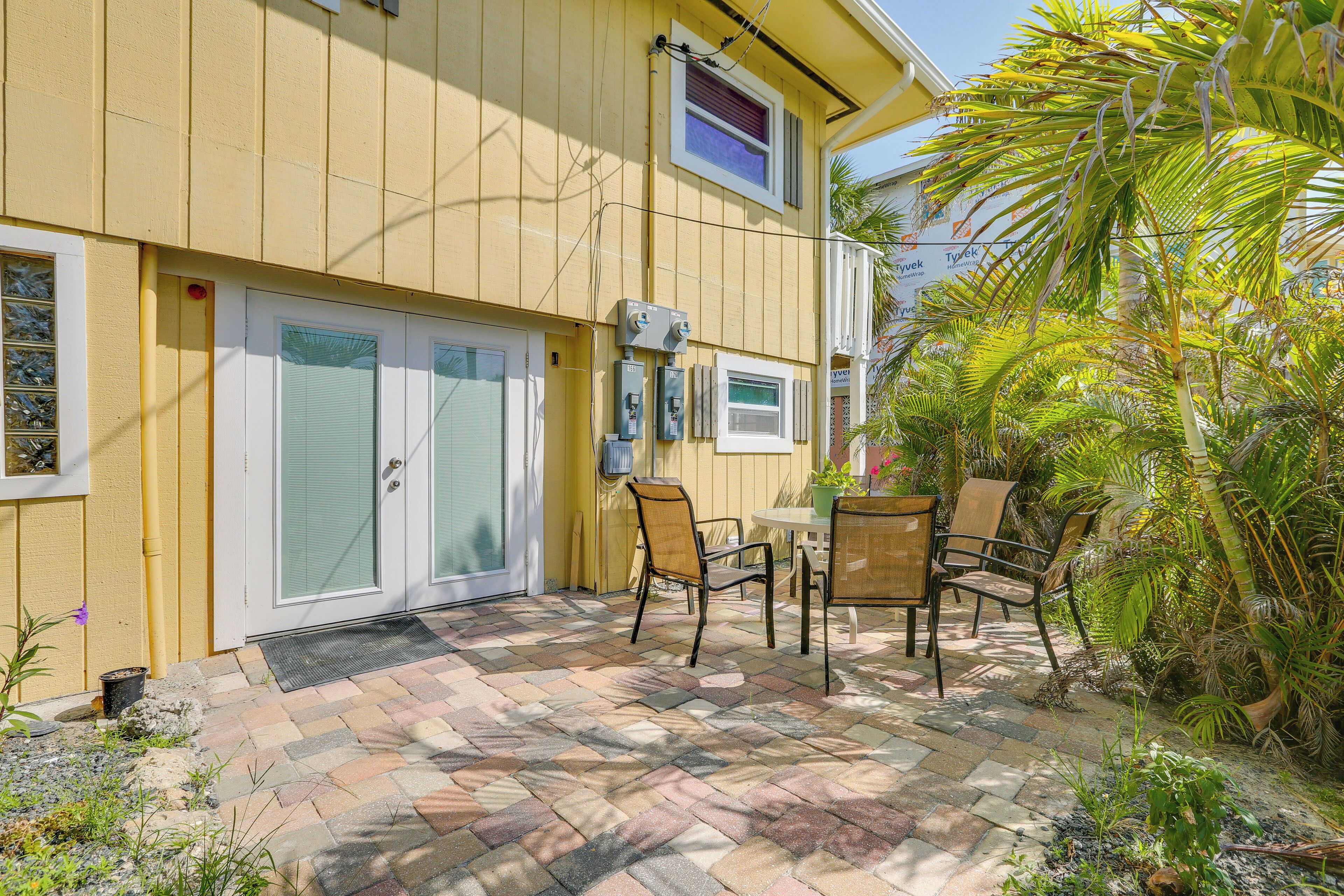 A yellow building with a covered patio area featuring a table and chairs, surrounded by potted plants and a palm tree.