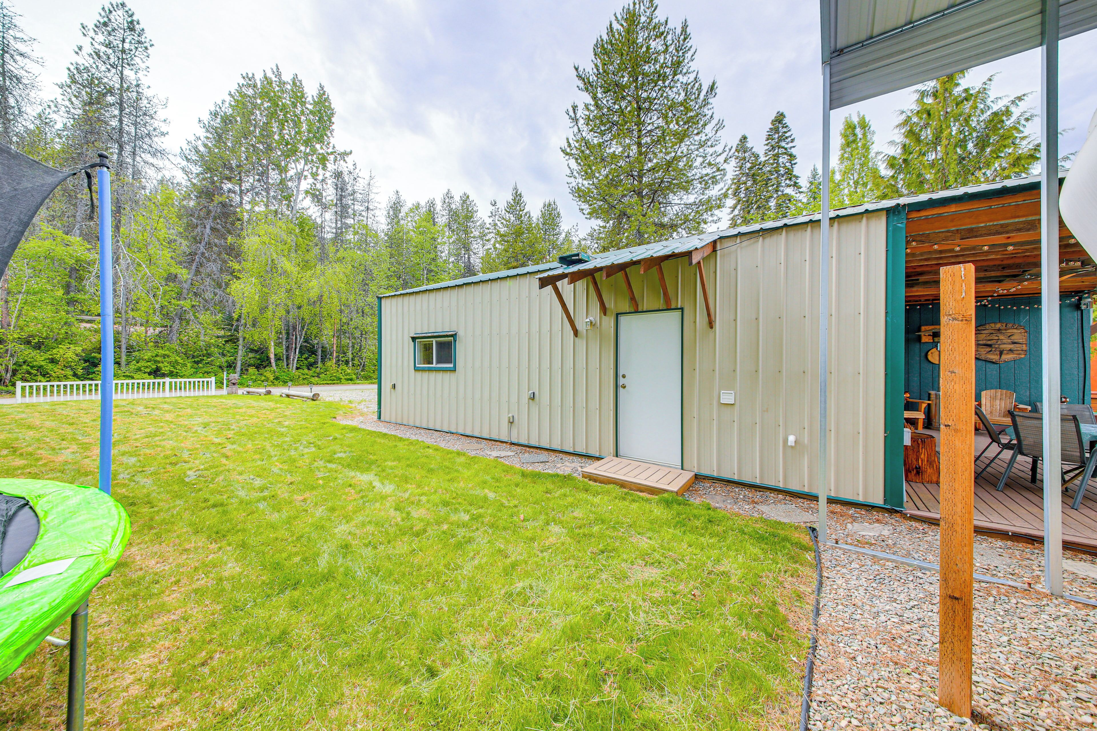 A metal building with a door and windows, a grassy yard, a deck with chairs, and a tree line in the background.