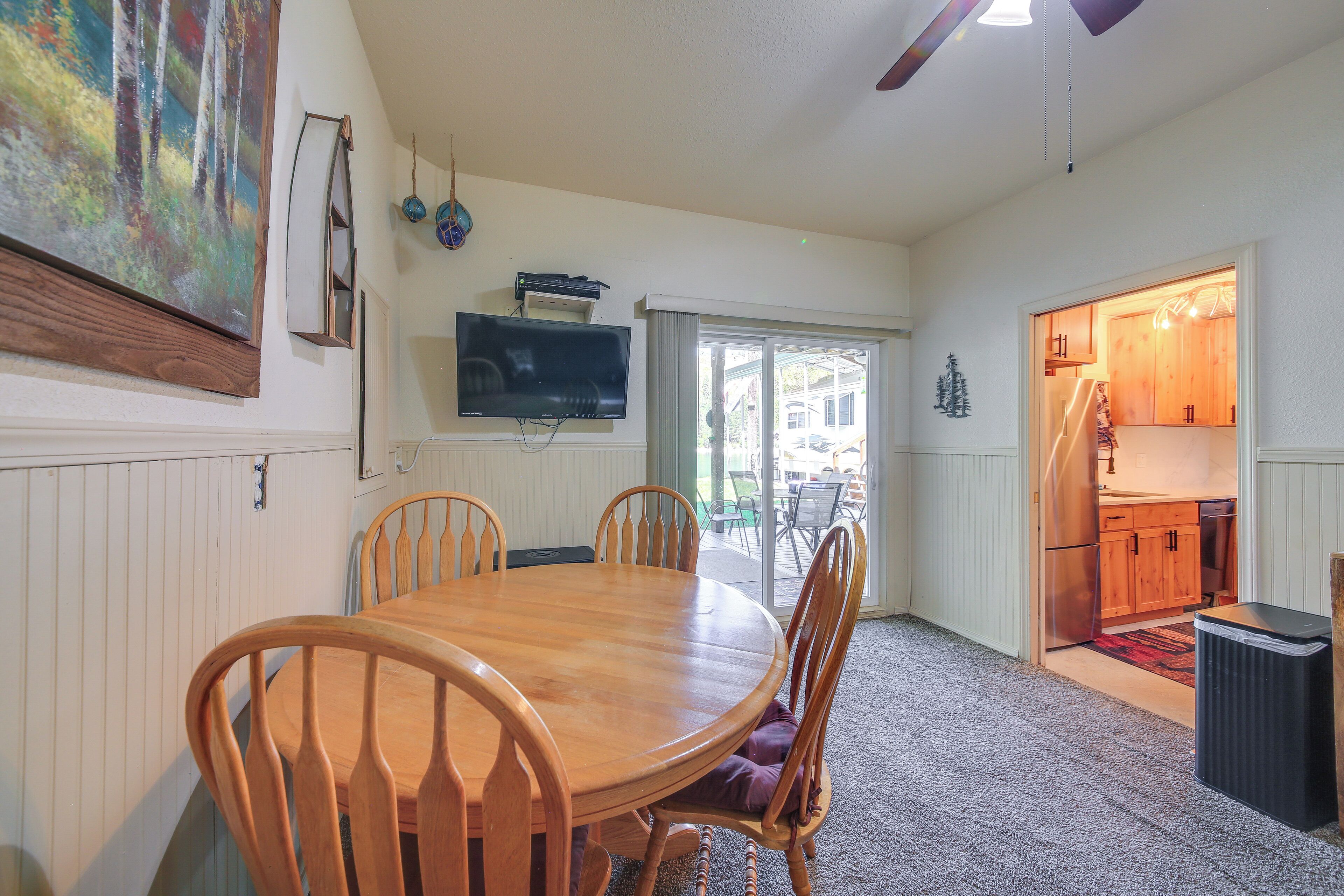 A dining area with a wooden table and chairs, a television mounted on the wall, and a kitchen visible through an open door.