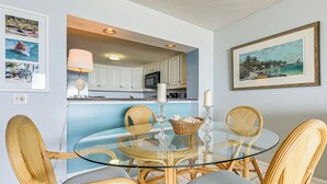 A dining area with a glass table surrounded by wicker chairs, a kitchen in the background, and framed artwork on the walls. - Oceanfront Resort Condo w/ Pools in Myrtle Beach (Myrtle Beach)