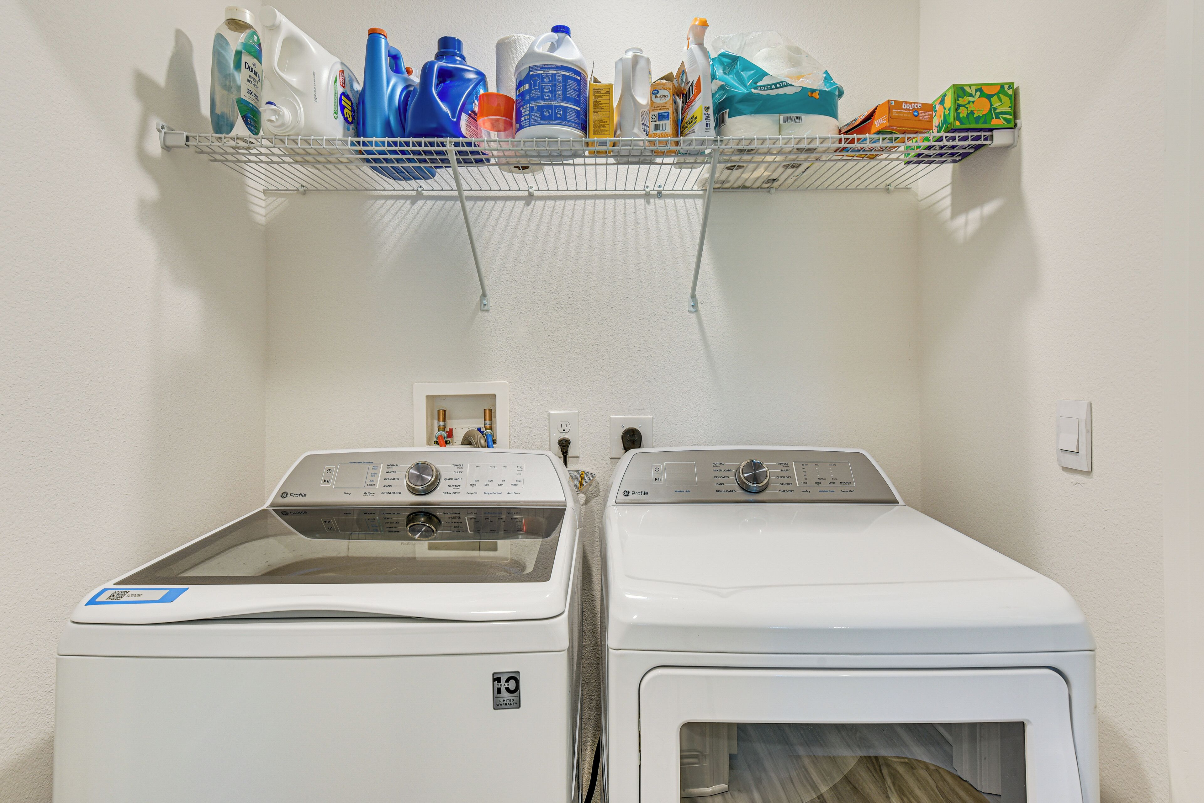 A laundry area with two front-loading washing machines and a shelf above containing various cleaning supplies.