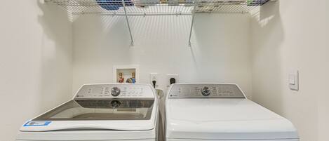 A laundry area with two front-loading washing machines and a shelf above containing various cleaning supplies.
