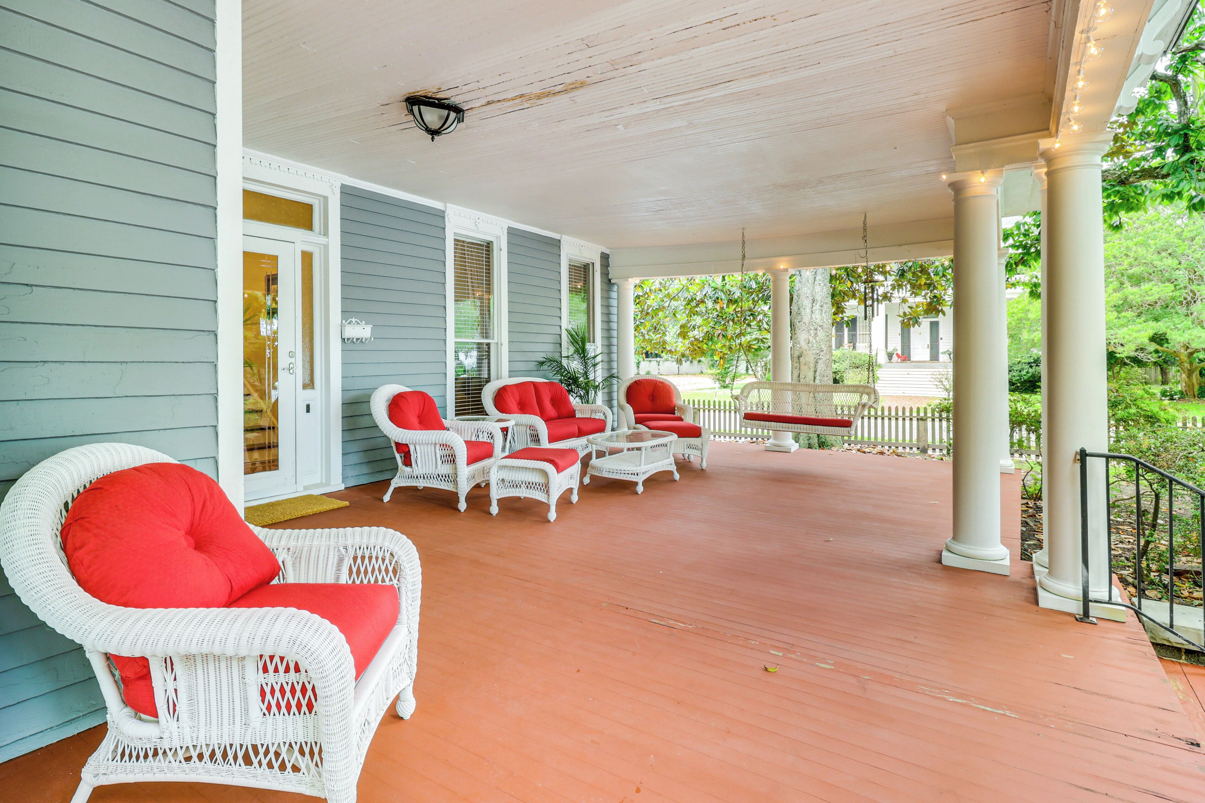 A covered porch with white wicker furniture and red cushions, a wooden floor, and a white picket fence in the background.