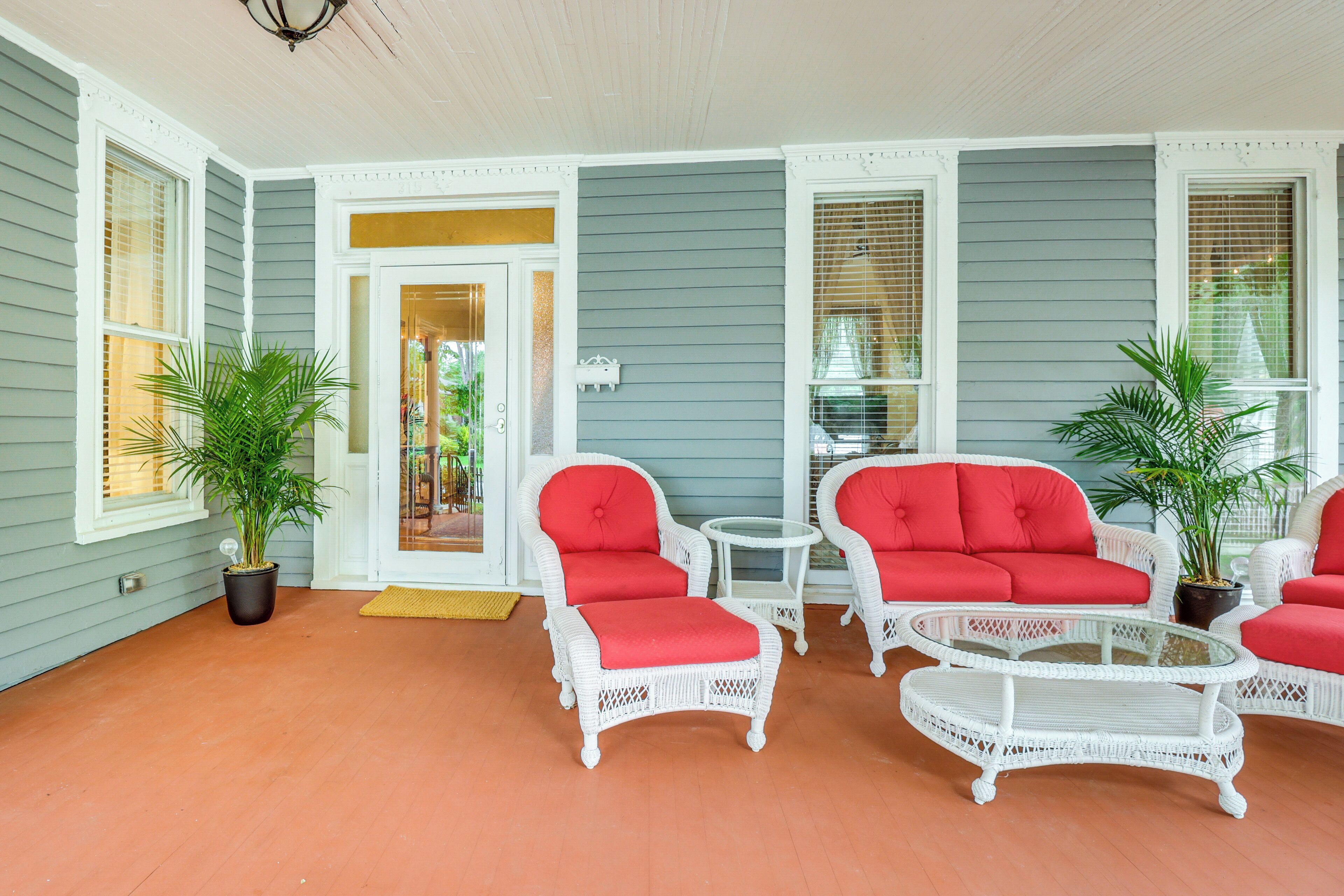 A covered porch with white wicker furniture and red cushions, a small table, and potted plants.