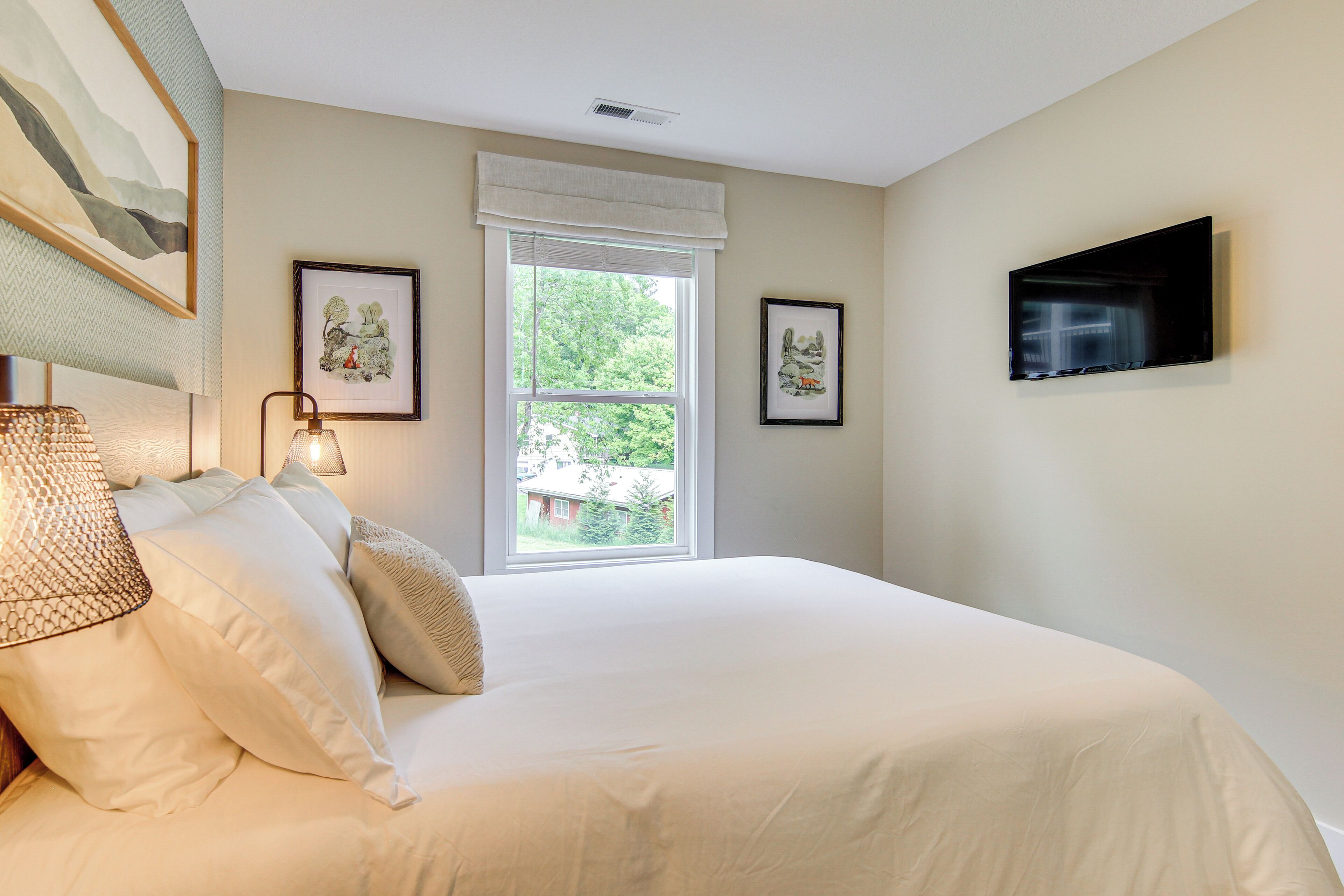 A bedroom with a large bed, a window with a view of greenery, and a flat-screen TV mounted on the wall.